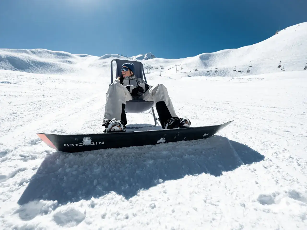 Snowboarder d&eacute;tendu sur la neige, se demandant s'il faut farter des skis neufs. Le soleil brille sur les pistes.
