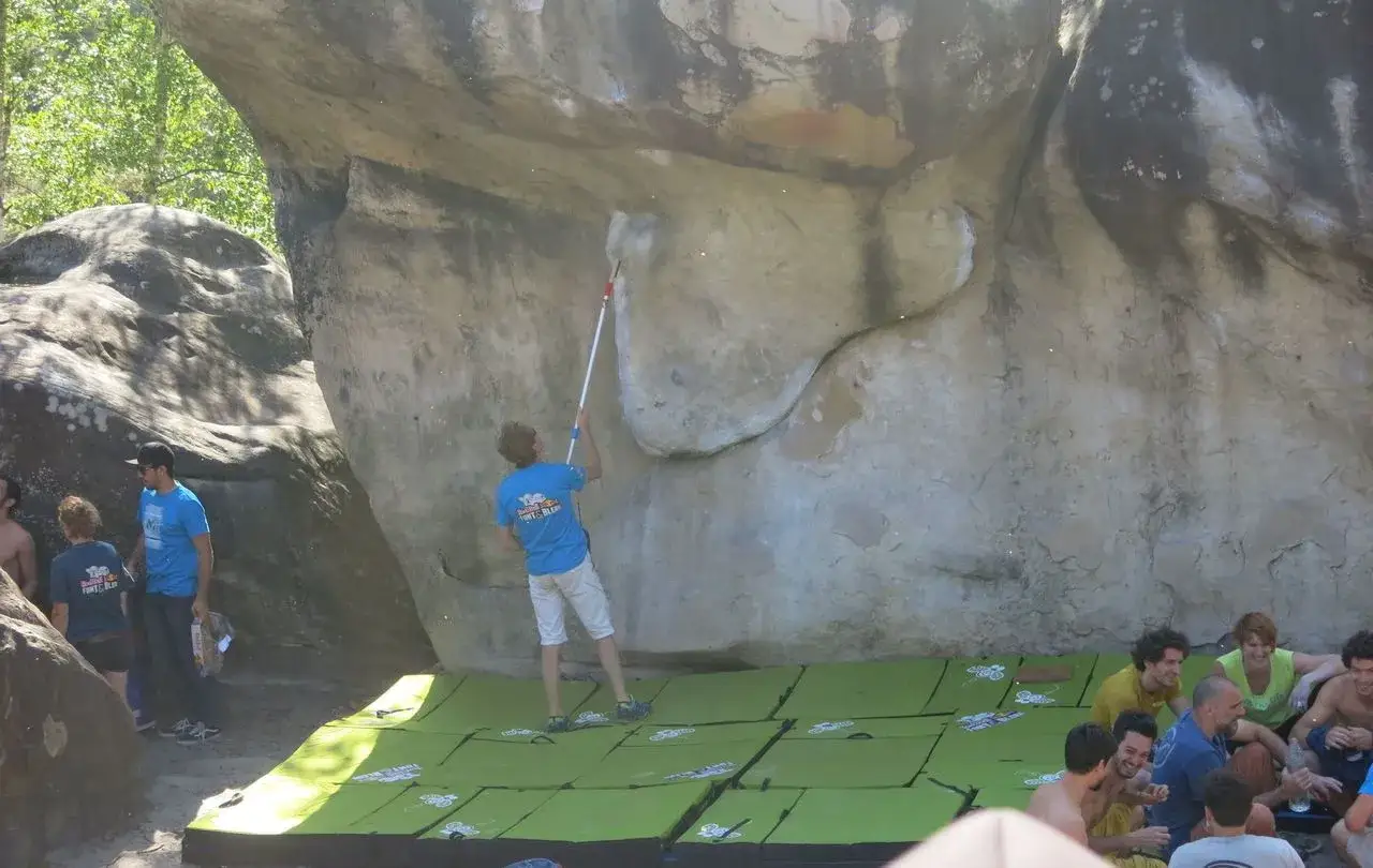 Un jeune homme nettoie un bloc escalade avec une brosse. Des grimpeurs attendent leur tour, assis sur des crashpads verts.