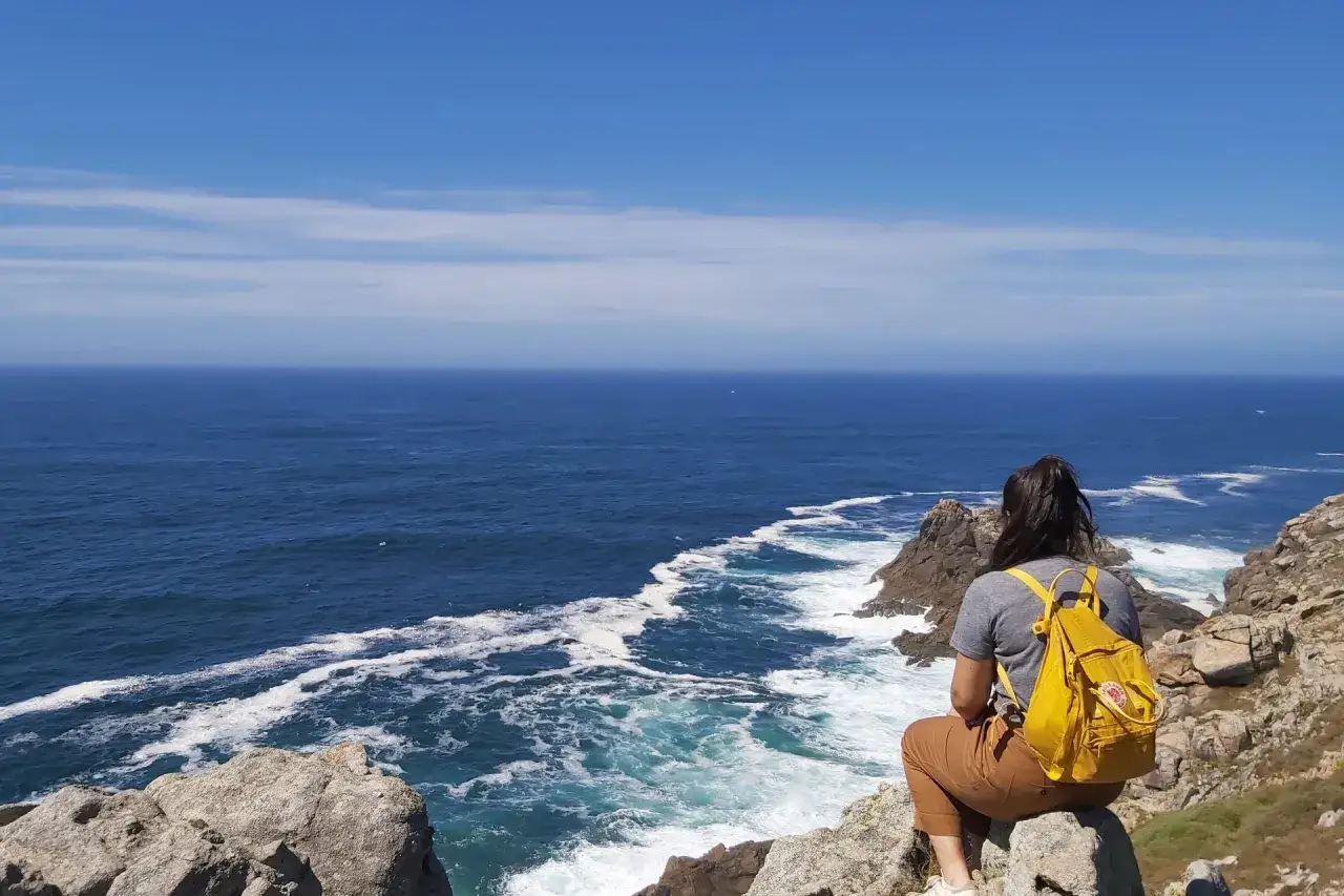Vue sur l'océan depuis le Camino dos Faros. Une randonneuse, sac jaune sur le dos, contemple les vagues déferlant sur la côte rocheuse.