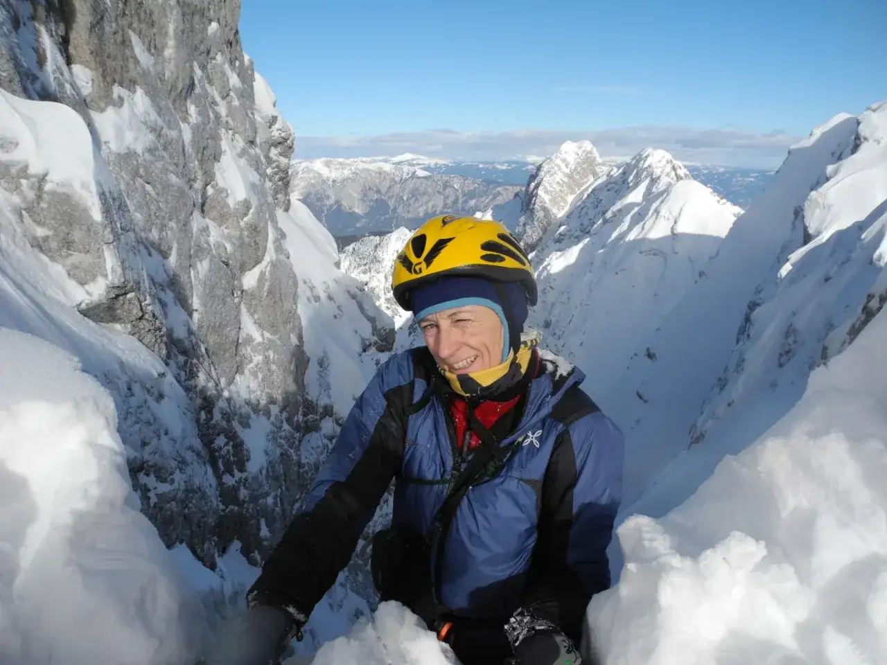 Une alpiniste souriante, Nives Meroi, en casque jaune, &eacute;volue dans un couloir enneig&eacute;, entour&eacute;e de sommets majestueux sous un ciel bleu.