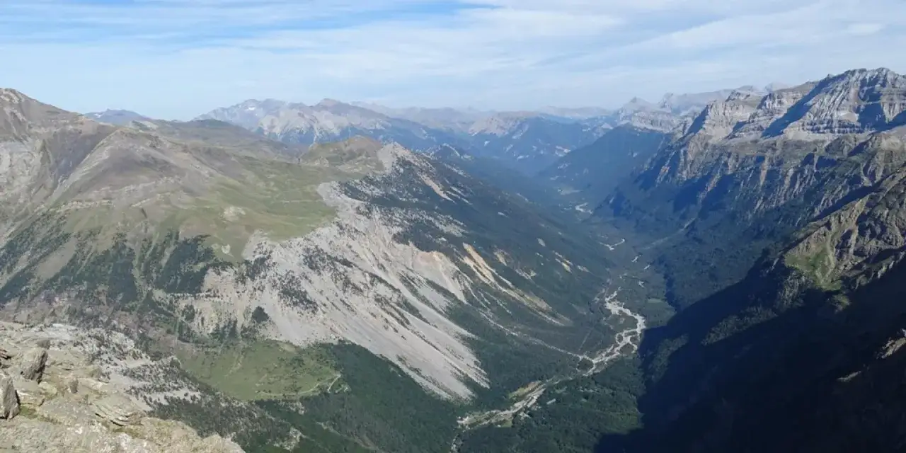 Vue panoramique des llanos de la Larri, une vallée verdoyante bordée de montagnes escarpées sous un ciel bleu.