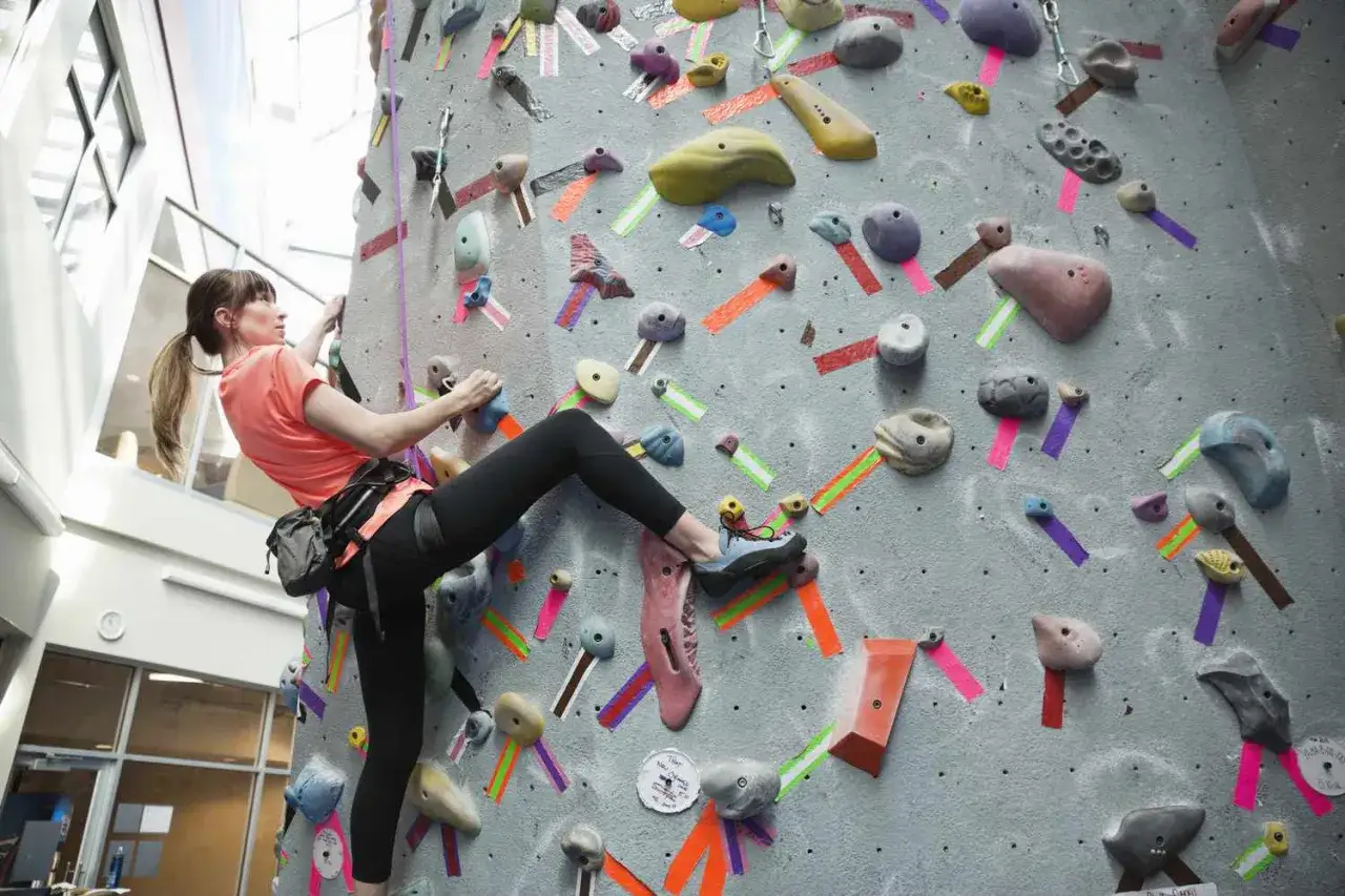Une femme fait de l'escalade, elle utilise des prises colorées pour grimper. Elle est concentrée sur son ascension.