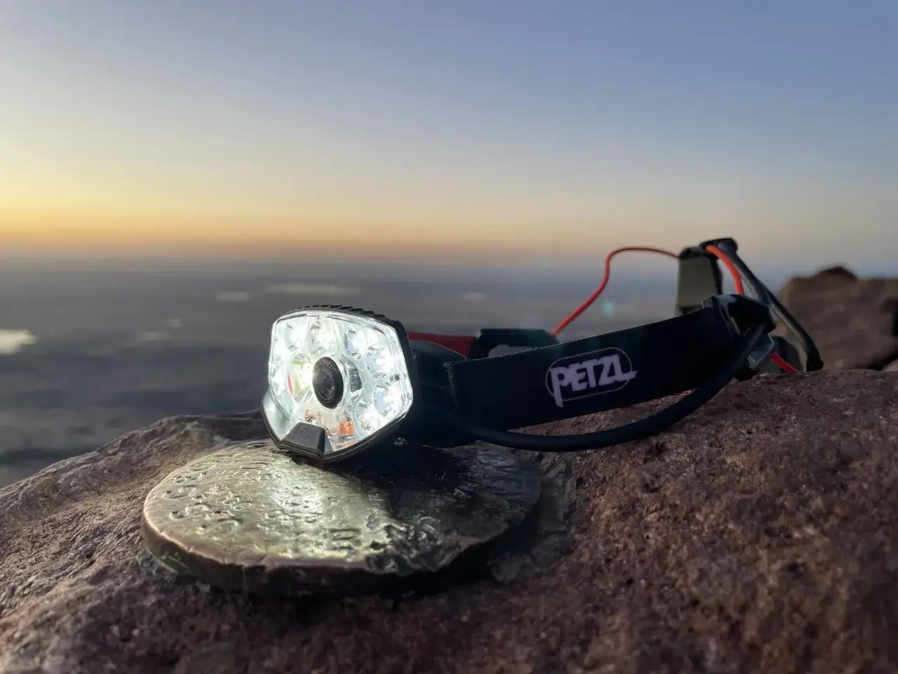 Lampe frontale Petzl Nao RL allumée sur un sommet rocheux au lever du soleil, prête pour un test d'aventure.