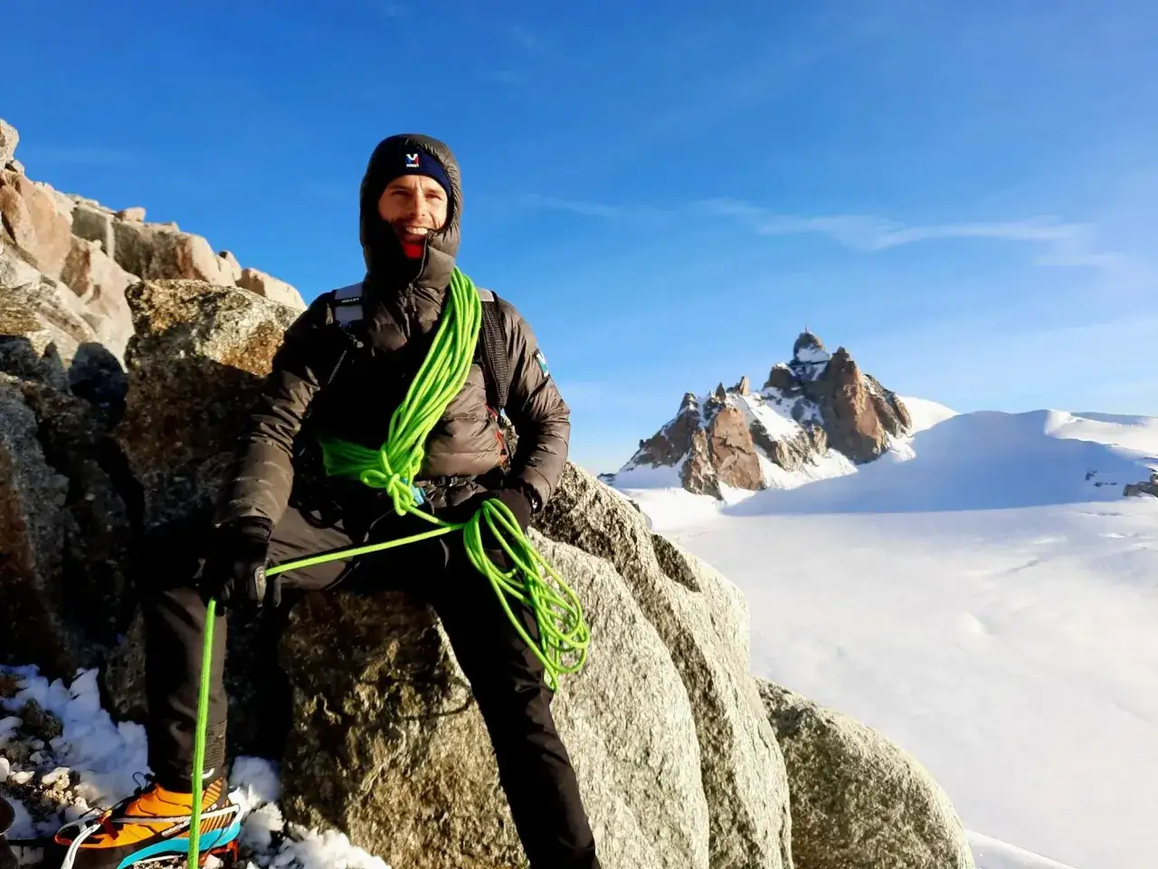 Un alpiniste souriant, corde verte enroul&eacute;e, sur un rocher enneig&eacute;. Le d&eacute;cor parfait pour un podcast alpinisme, avec des sommets enneig&eacute;s en arri&egrave;re-plan.