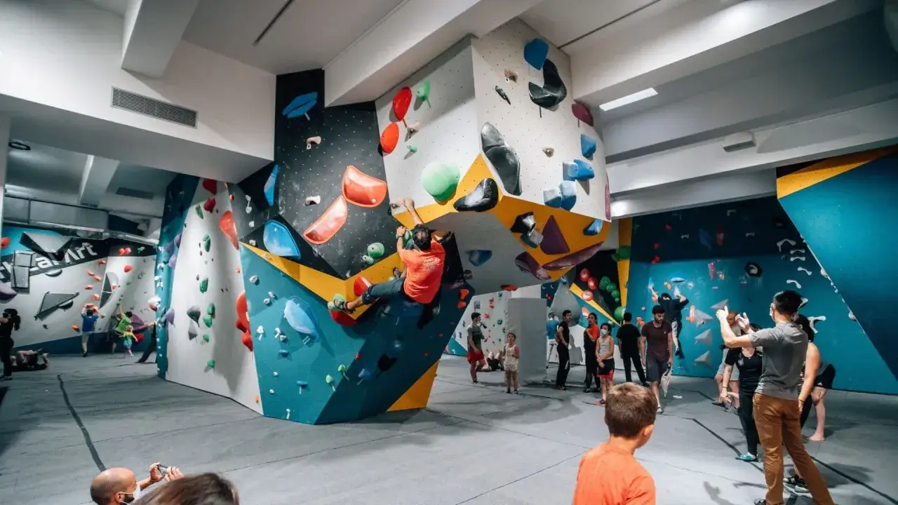 Un homme en t-shirt orange pratique l'escalade sur un mur color&eacute;, entour&eacute; d'autres grimpeurs et spectateurs dans une salle d'escalade. C'est un vrai centre d'escalade nation !