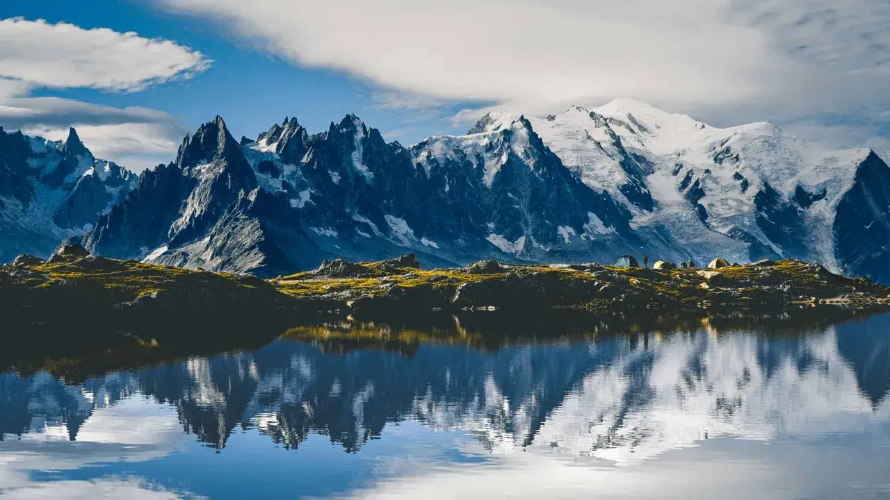 Un lac reflète un majestueux sommet montagneux enneigé, avec des tentes de campeurs sur la rive rocheuse.