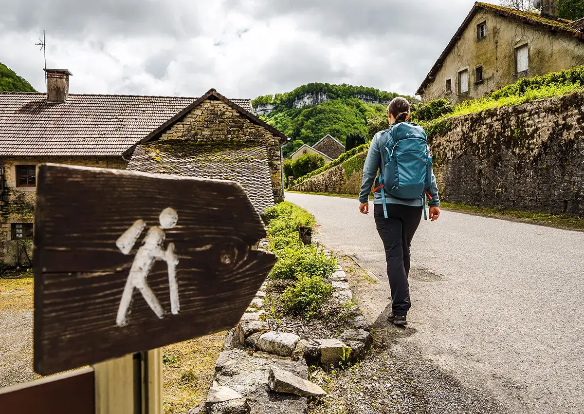 Panneau de randonnée indiquant la direction. Une femme avec un sac à dos bleu entame son premier trek dans un village pittoresque.