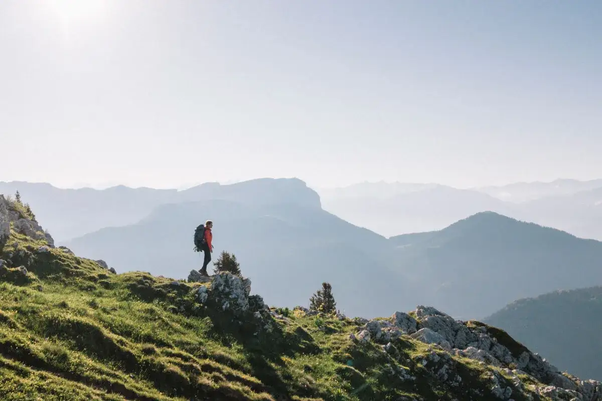 Une personne randonne seule sur une crête herbeuse, contemplant des montagnes bleutées à perte de vue.