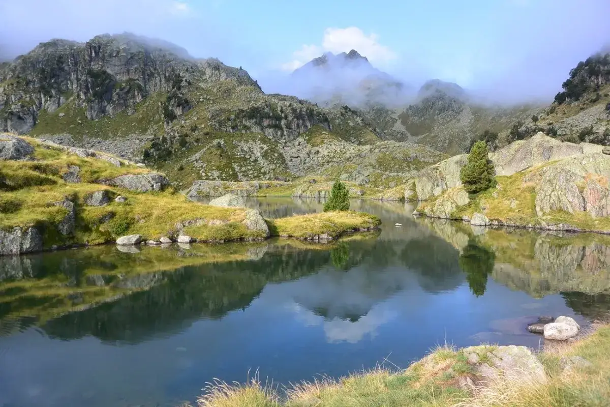 Calme lac refl&eacute;tant les montagnes rocheuses et verdoyantes du cirque de Colomers, avec un ciel bleu et des nuages bas.