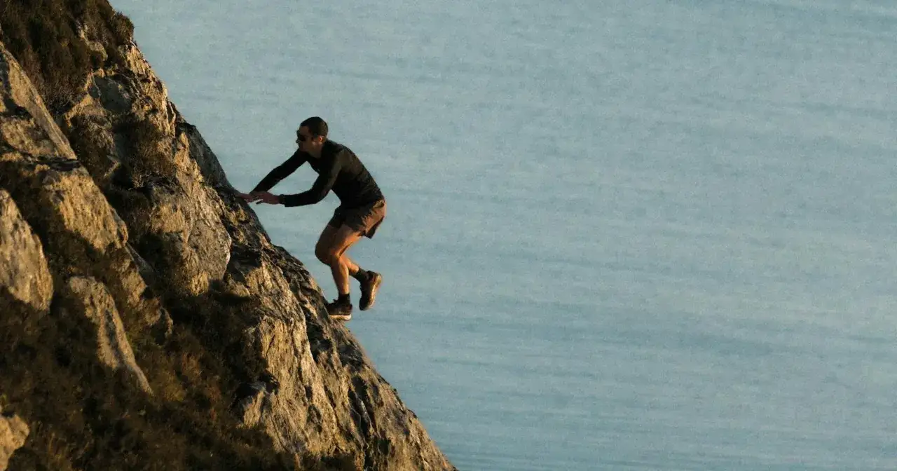 Kilian Jornet escalade une paroi rocheuse abrupte, dominant un plan d'eau calme. Une sc&egrave;ne typique de ses aventures dans les Pyr&eacute;n&eacute;es.