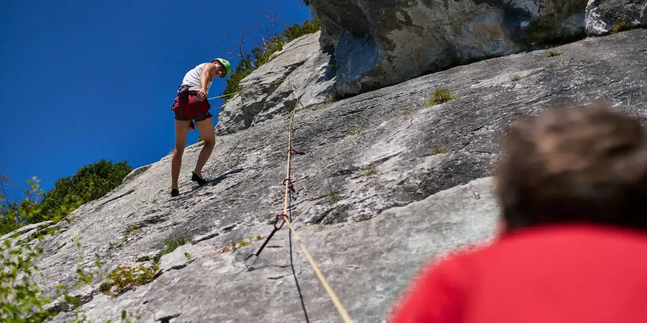 Un grimpeur en plein effort d'escalade 64, harnach&eacute; et casqu&eacute;, progresse sur une paroi rocheuse sous un ciel bleu &eacute;clatant.