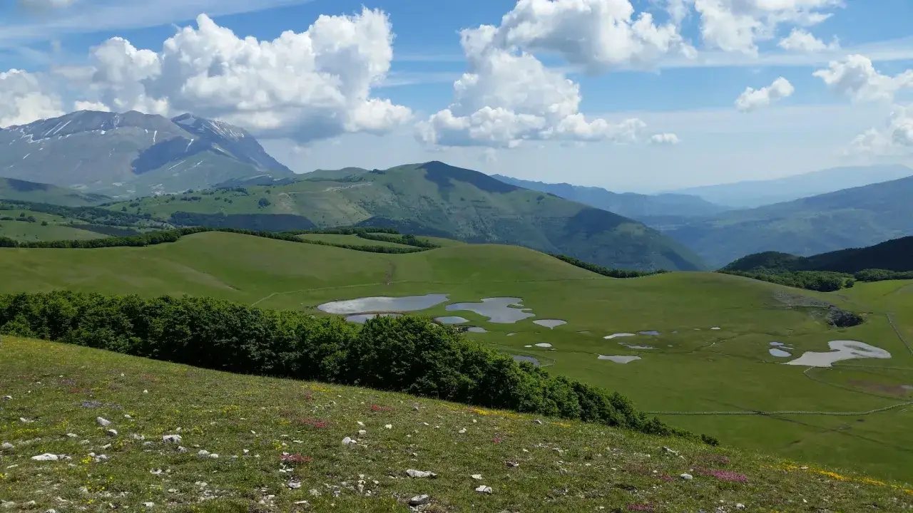 Vastes prairies verdoyantes parsemées de petits étangs, avec des montagnes majestueuses en arrière-plan, typiques du parc national des monts Sibyllins.