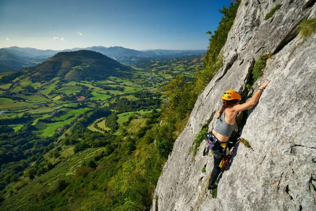 Une femme en casque orange pratique l'escalade dans le Lot, dominant un paysage verdoyant de collines et de champs.