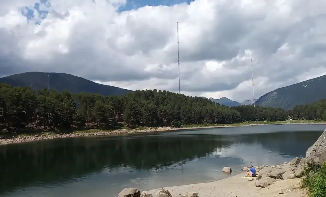 Vue paisible du lac d'Engolasters, entour&eacute; de for&ecirc;ts de pins et de montagnes. Une personne assise sur la rive profite du calme.