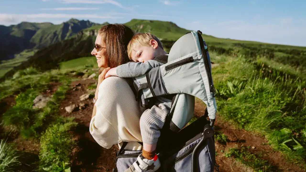 Une m&egrave;re souriante porte son jeune enfant endormi dans un porte b&eacute;b&eacute; rando age, sur fond de montagnes verdoyantes.