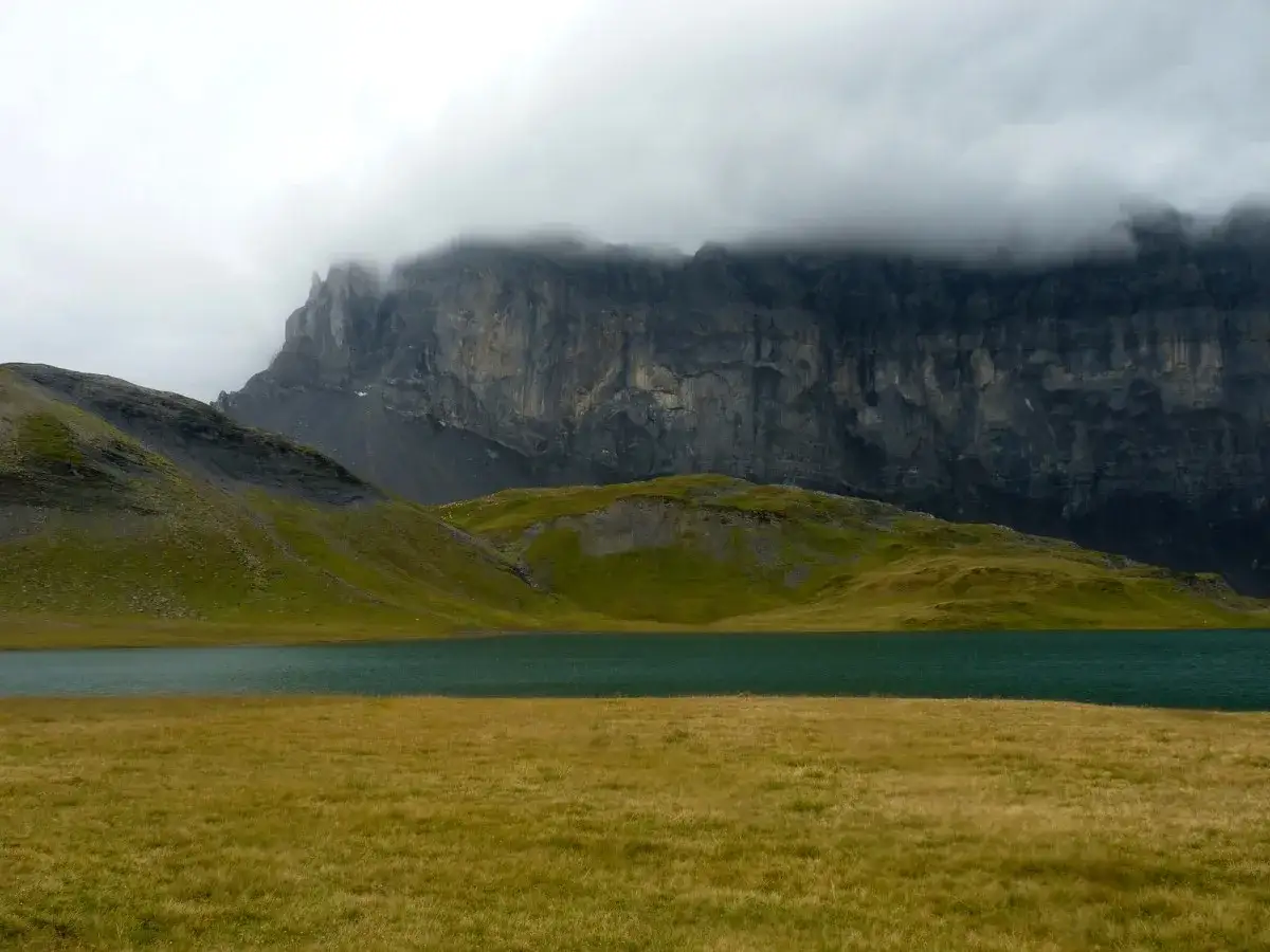 Vaste paysage alpin avec un lac turquoise, des collines verdoyantes et une imposante paroi rocheuse voil&eacute;e de nuages, &eacute;voquant le tour des Fiz.