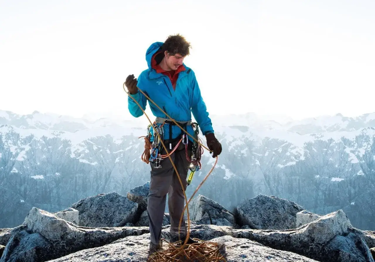 Grimpeur en veste shell, c'est quoi ? Il manipule une corde sur un sommet rocheux, avec des montagnes enneig&eacute;es en arri&egrave;re-plan.