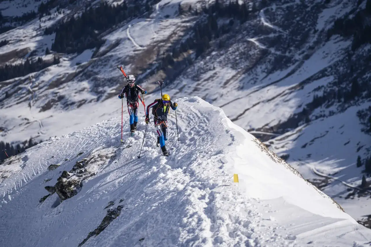 Deux skieurs escaladent une crête enneigée, leurs skis sur le dos. Ils utilisent des bâtons pour s'aider, comme s'ils étaient sur une peau de phoque, prêts pour la descente.