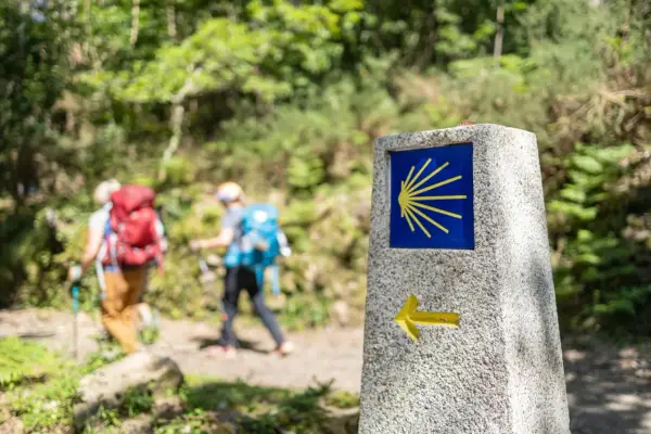 Panneau du Camino de Santiago avec une coquille jaune sur fond bleu et une flèche. Deux randonneurs avancent sur le chemin.