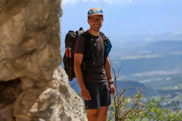 Jérémy Bigé, souriant, prêt pour l'aventure avec son sac à dos et ses bâtons de randonnée, dominant un paysage de montagnes.